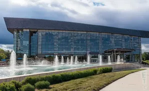 A modern building with a glass facade and a dark, angled roof, featuring a large fountain in front.