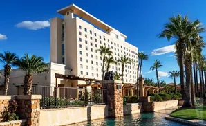 A tall, beige hotel with many windows stands beside a canal and palm trees.