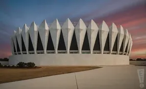 A modern, white, circular building with pointed arches against a colorful sunset sky.