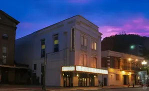 A historic theater with a lit marquee stands on a street at twilight, with a purple and blue sky above.