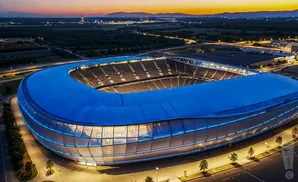 Aerial view of a modern stadium at dusk, illuminated with blue lights, with a crowd in the stands.