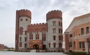 Castle-like brick building with two towers and a connecting section, adjacent to a modern tan building.