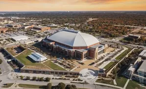 Aerial view of a large domed stadium with surrounding buildings and trees under a warm, orange sky.