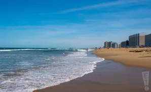 A wide beach with ocean waves lapping the shore, backed by hotels under a bright blue sky.