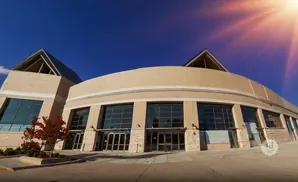 Wide shot of a tan building with large glass windows, under a bright blue sky with sun flares.