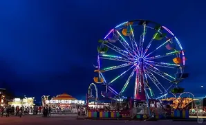 A Ferris wheel and carousel at a carnival at night, lit by colorful lights.