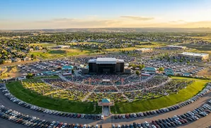 An aerial view of an outdoor concert venue with a large crowd, stage, and surrounding parking lots at sunset.