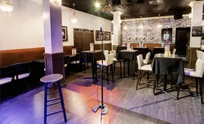 A comedy club stage with tables, chairs, and a microphone stand on a wooden floor.