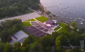 Aerial view of an outdoor amphitheater with a stage and audience, located by a body of water with many boats.