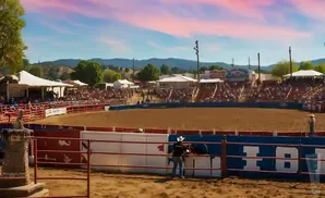 Rodeo arena with spectators, cowboys, and a bull under a colorful sunset sky.