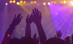 Silhouetted hands raised in a concert venue bathed in purple and yellow stage lights.