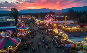 Aerial view of a bustling fair at sunset, with colorful rides and a crowd of people under a vibrant sky.