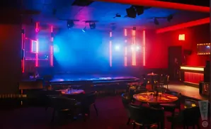 A nightclub with a stage, tables, chairs, and a bar, lit by red and blue neon lights.