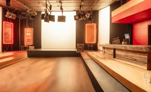 A stage area with a wooden desk, microphones, and a screen, set against a backdrop of black curtains and a large white screen.