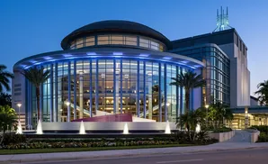 A modern theater building with a glass facade and dome, illuminated by blue lights at dusk.