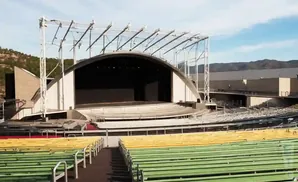 Outdoor amphitheater with tiered seating in green, yellow, and red, facing a large stage with a curved roof structure.
