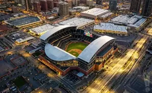 Aerial view of a baseball stadium at dusk, with a lit field and surrounding urban landscape.