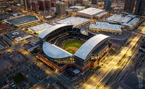 Aerial view of a baseball stadium at dusk, with city buildings and lights in the background.