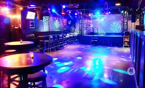 A nightclub stage with a screen, bar, tables, and dance floor lit with blue and purple lights.