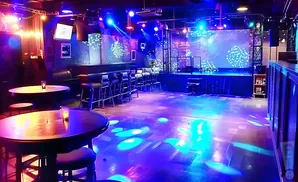 A dimly lit club with a stage, bar, tables, and colorful lights projected onto the floor.