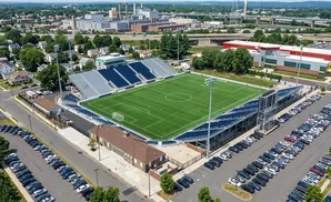 Aerial view of a soccer stadium with a green field, bleacher seating, and surrounding parking lots.