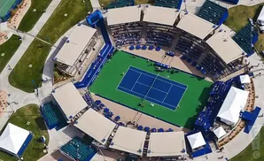 Aerial view of a tennis court with tiered seating and shaded canopies.