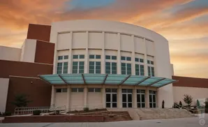 Modern building with a curved facade, large windows, and a glass canopy over the entrance, under a dramatic sunset sky.