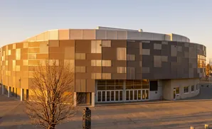 A modern, curved building with a patterned facade of metal panels in warm tones and gray, reflecting the sunset.