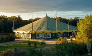 A large, green circus tent with a yellow trim sits in a grassy field at sunset.