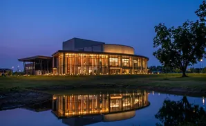 A modern building with large glass windows reflects in a dark pond at dusk.