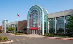 A modern shopping mall entrance with large glass arched windows, a red awning, and a flagpole.