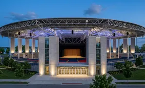 An open-air amphitheater with a curved roof, concrete pillars, and tiered seating under a twilight sky.