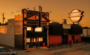 A retro diner with orange and black accents, featuring a tall illuminated sign and a circular sign at sunset.