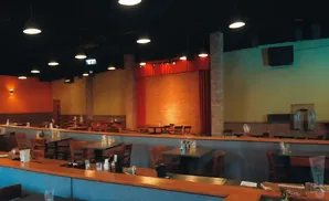 Interior of a restaurant with tables and chairs, a brick stage area with red curtains, and orange and yellow walls.