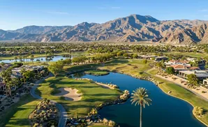 Aerial view of a luxurious golf course with a lake, palm trees, and mountains in the background.