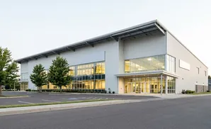 A modern, light-colored industrial building with large windows and a gray metal roof sits next to a parking lot.