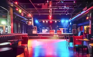 Empty concert venue with stage, drum set, amps, and seating, illuminated by colorful stage lights.