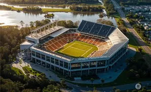 Aerial view of a stadium with orange seats and a green field, surrounded by trees and water.