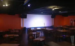 A dimly lit comedy club with orange walls, a large white stage, tables, and chairs.