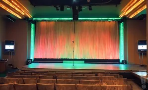A stage with a microphone, curtains lit with red and green lights, and rows of empty seats.