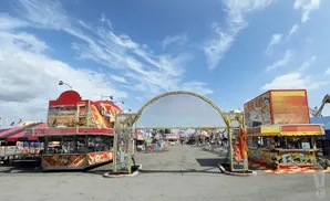 Entrance archway to a carnival midway with food stands and rides under a blue sky with clouds.