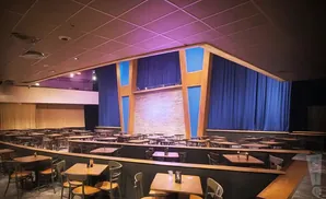 Comedy club stage with brick backdrop, blue curtains, and rows of tables and chairs.