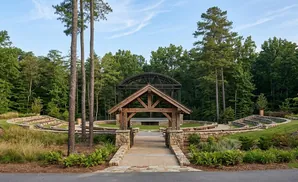 An outdoor amphitheater with wooden archway, stone seating, and a covered stage in a forest setting.