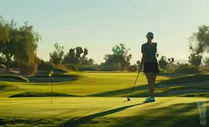 Golfer on a sunny course, preparing to putt, with a flag and rolling green hills in the background.