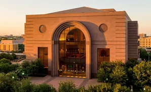 Large tan building with a grand arched entryway and windows, surrounded by lush green trees under a warm sunset sky.