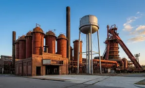 An abandoned industrial complex with tall red silos, a smokestack, and a water tower under a blue sky.