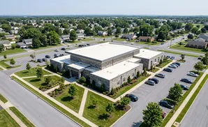 Aerial view of a modern stone-clad building with a flat white roof, surrounded by a parking lot and lush green landscaping, with a suburban neighborhood…