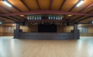 Empty ballroom with a stage, wooden floor, and ceiling beams.