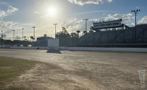 Sunlight shines on a dirt racetrack with grandstands in the background under a cloudy sky.