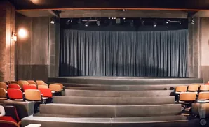 An empty theater with tiered seating facing a stage with blue curtains.